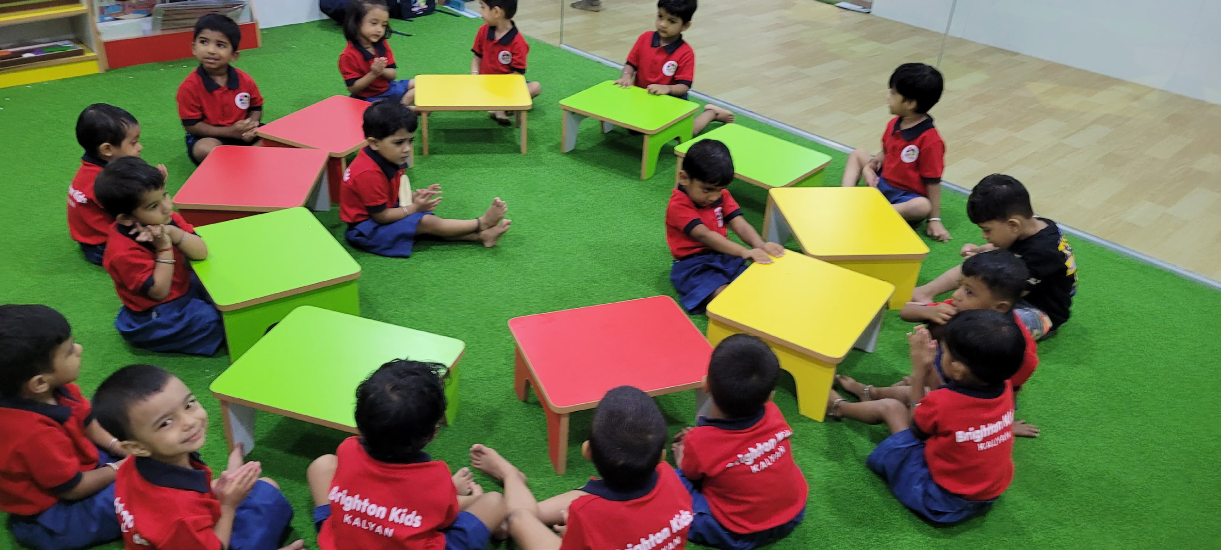 Group of preschool children sitting in a circle on green mat with colorful tables during classroom activity